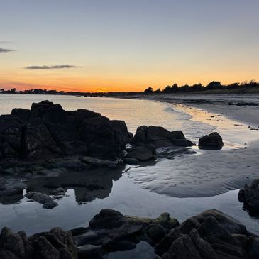 Sunset over a rocky beach with calm water and silhouetted trees.