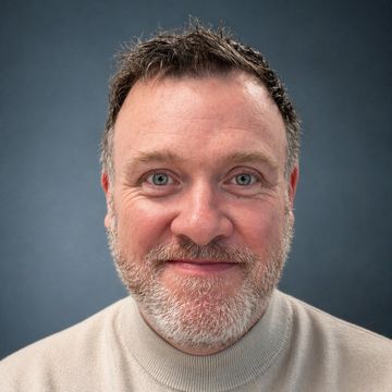 Smiling middle-aged man with a trimmed beard and light sweater against a gray background.