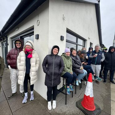 People dressed warmly standing and sitting outside a building on a cloudy day.