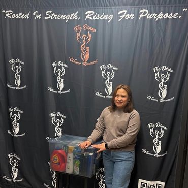 A woman stands smiling beside a donation box with a backdrop reading 'Rooted In Strength, Rising For Purpose.'