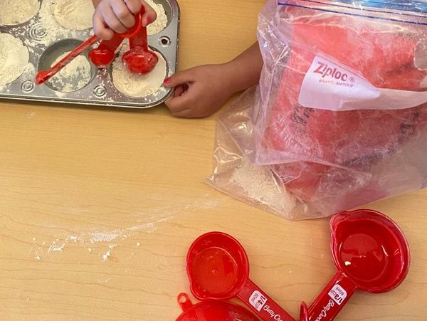 Child scoops flour into muffin tin using red measuring cups.