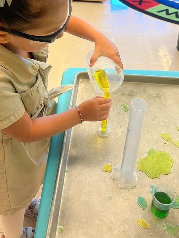 Child wearing safety goggles pouring yellow liquid into a measuring cylinder during a science experiment.
