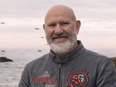 Smiling man with a beard wearing a leather jacket by the sea.