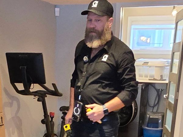 Man with a beard and black outfit standing near exercise bike indoors.