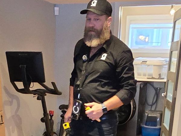 Man with a beard and cap standing near exercise bike indoors.