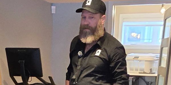 Man with a beard and black uniform stands near exercise equipment indoors.