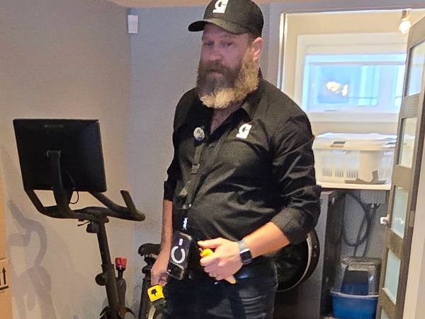 Bearded man in black shirt and cap holding tools in a room with exercise bike.