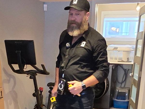 A bearded man in black uniform stands near an exercise bike indoors.