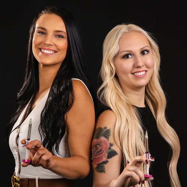 Two women smiling, holding syringes, one with black hair and the other with blonde hair and a rose tattoo.