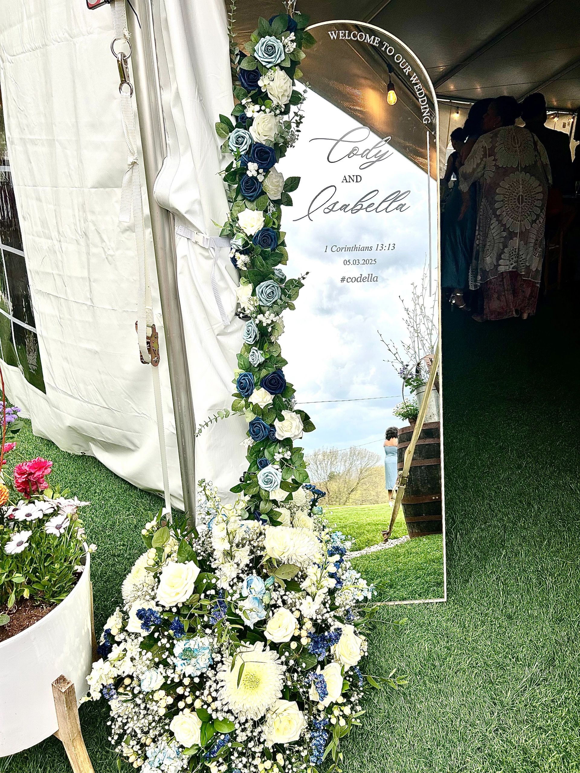 Elegant wedding mirror sign with flowers and guests reflected inside tent.