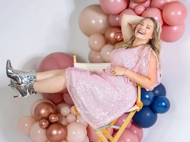 A joyful woman in a pink sequined dress and silver boots lounges on a chair surrounded by colorful balloons.