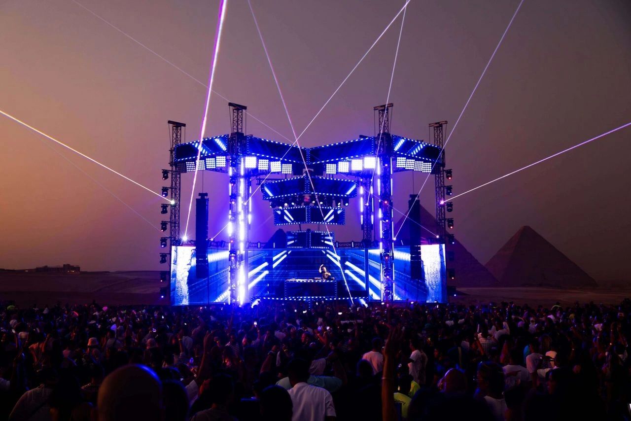 A DJ on stage before a huge crowd at the foot of the pyramids of Giza, lit up at dusk during a Pyramids Sessions night