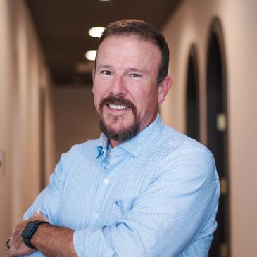 Smiling man with a beard in a light blue shirt stands confidently in a hallway.