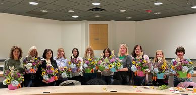 A group of women holding colorful flower arrangements, smiling indoors.