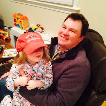 A man smiling while holding a young girl in a red cap indoors.