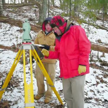 Two people conducting land surveying in a snowy forest.