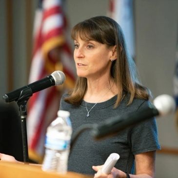 Woman speaking at a podium with American flag in the background.