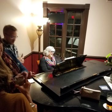 An elderly woman plays piano as others enjoy drinks nearby in a cozy room.