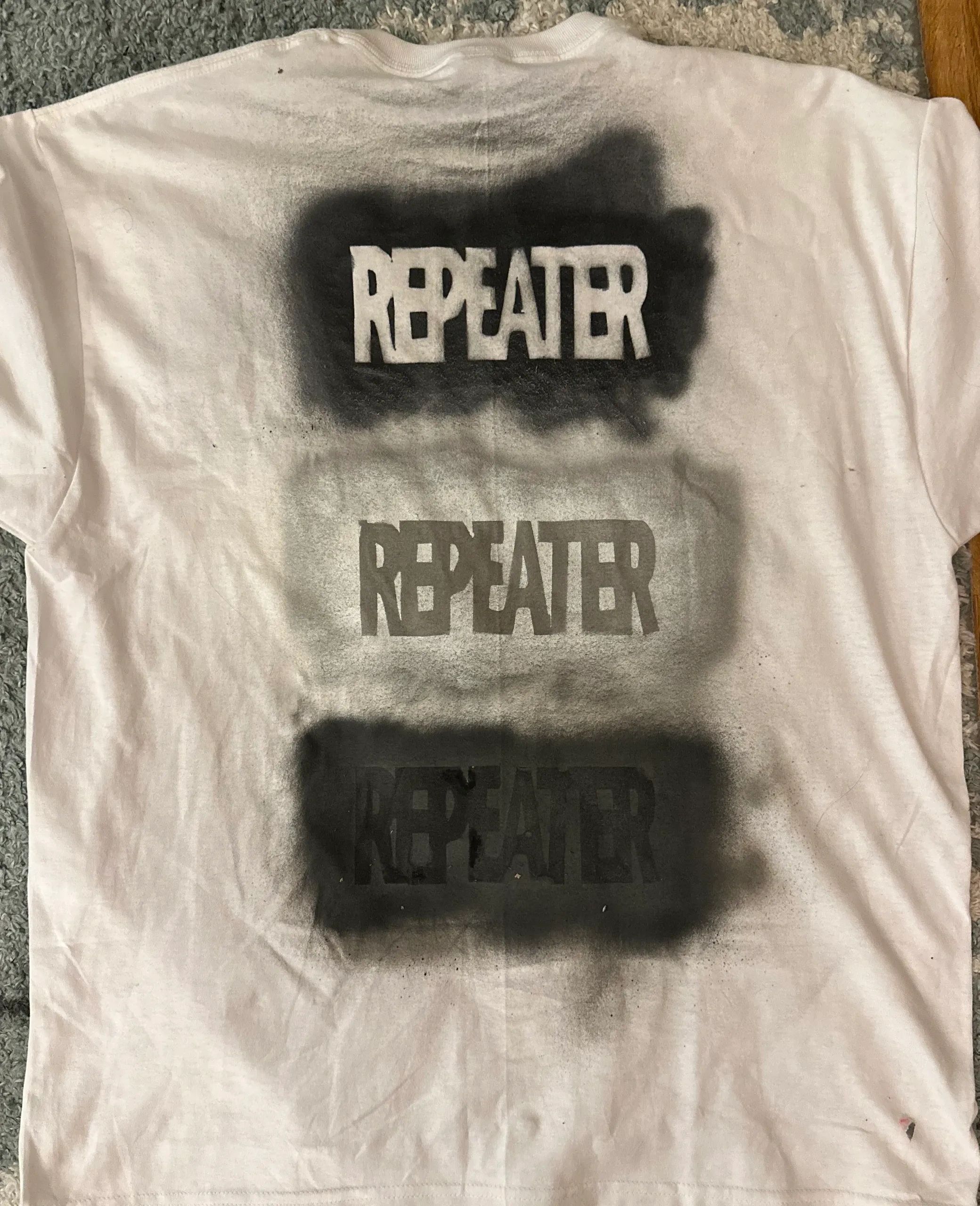 White shirt with the word 'REPEATER' spray painted three times in black.