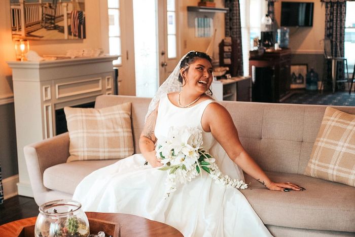 Bride in a white wedding dress smiling on a cozy couch indoors.