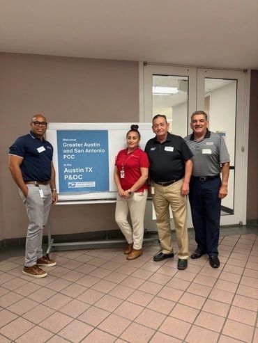Four people standing near a sign welcoming Austin and San Antonio PCC.