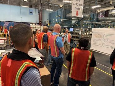 Workers in safety vests listening to a presenter in a warehouse.