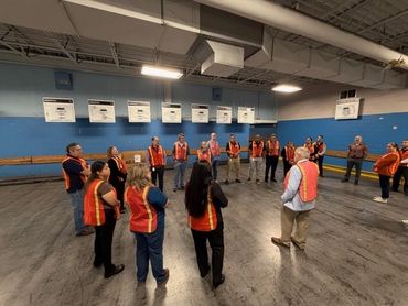 Group of people in orange safety vests standing in a circle indoors.