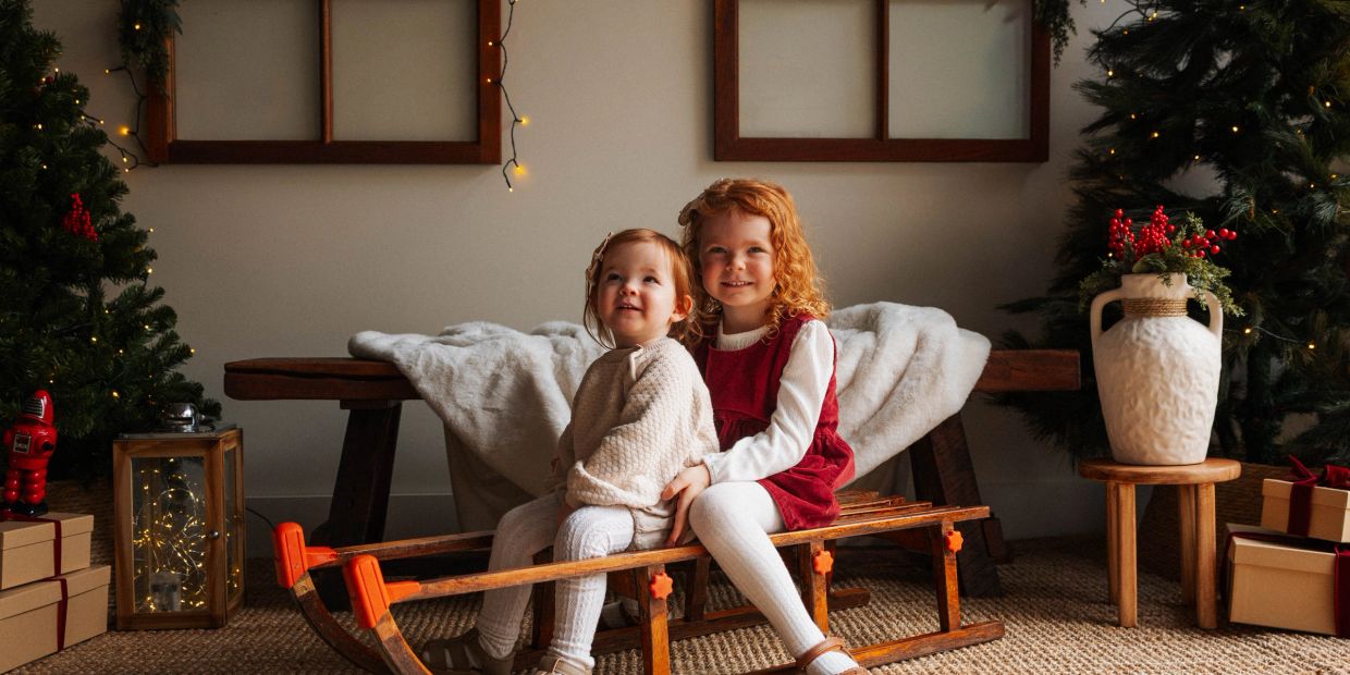 Two children smiling on a wooden sled indoors near Christmas decorations.