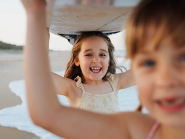 Two young sisters playing together at the beach, with one girl smiling brightly while holding a beac