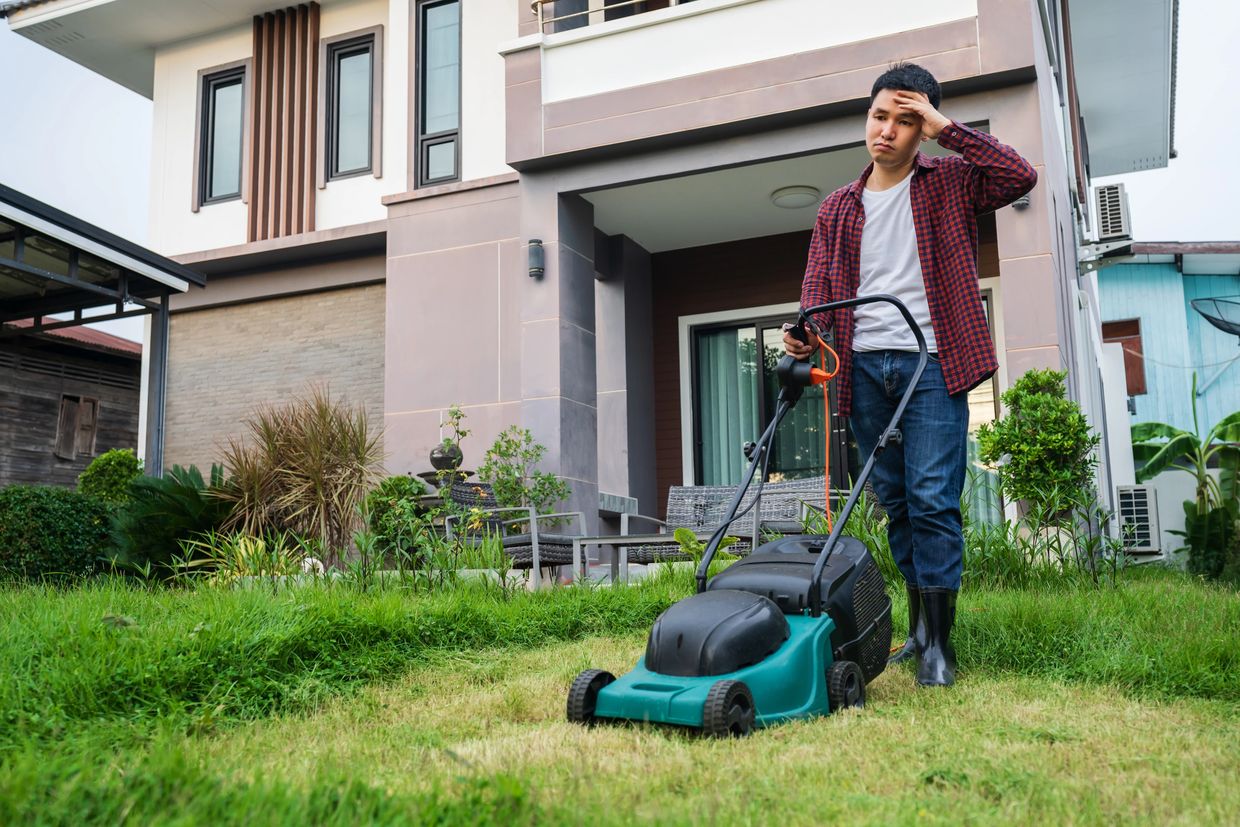 Man frustrated with lawn mower in front of house with overgrown grass.