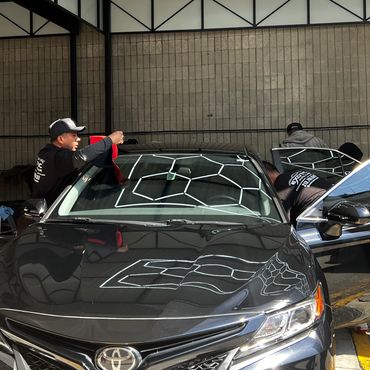 Two men cleaning a black Toyota car inside a garage with geometric ceiling lights.