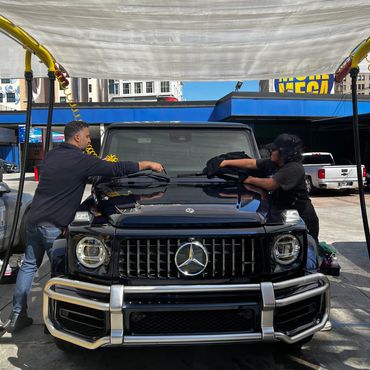 Two people cleaning the windshield of a black Mercedes-Benz G-Class SUV.