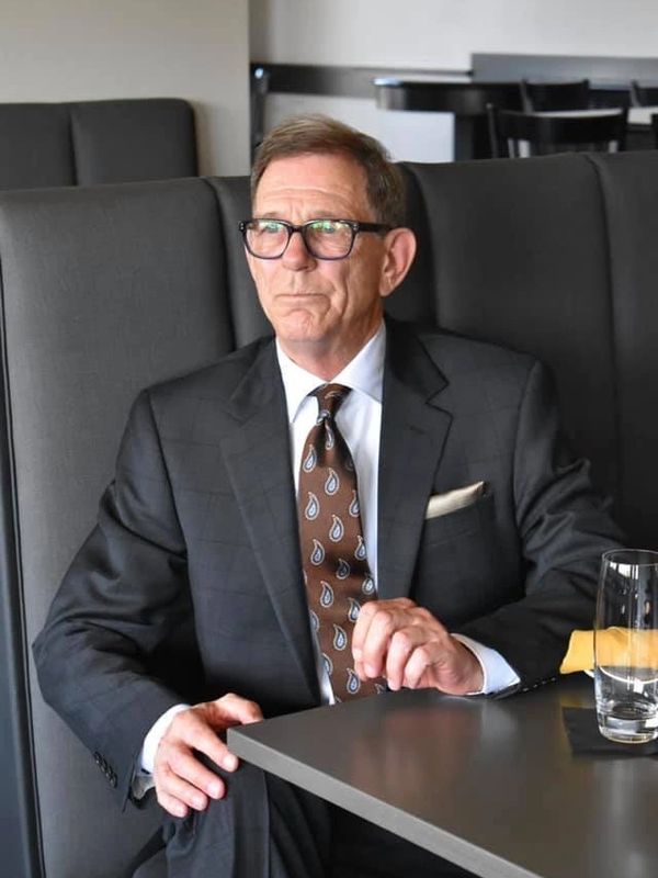 A man in a suit and glasses sitting at a table looking thoughtful.