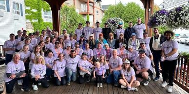 Large group of people wearing matching purple shirts posing on a wooden porch.