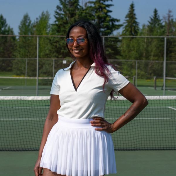 Woman posing on tennis court in white outfit with sunglasses.