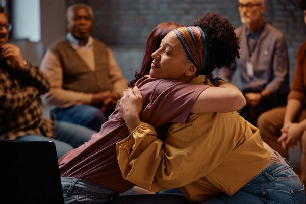 Two women embrace in a supportive group therapy session, with others blurred in the background.