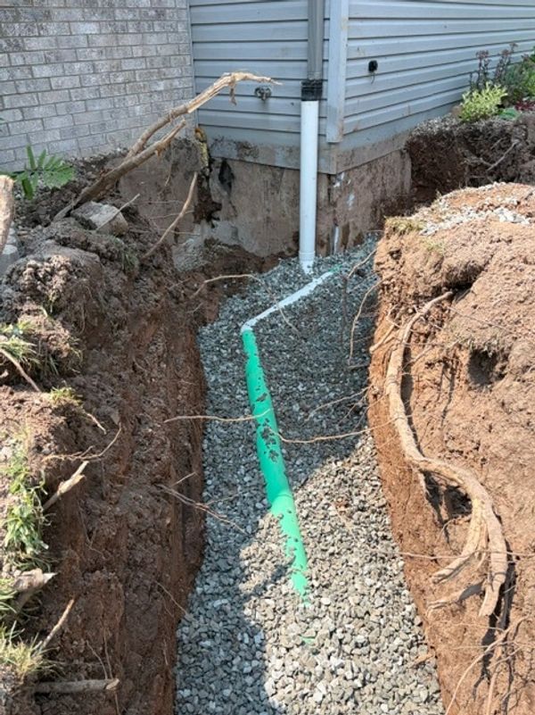 Trench dug for drainage pipe installation near house foundation.