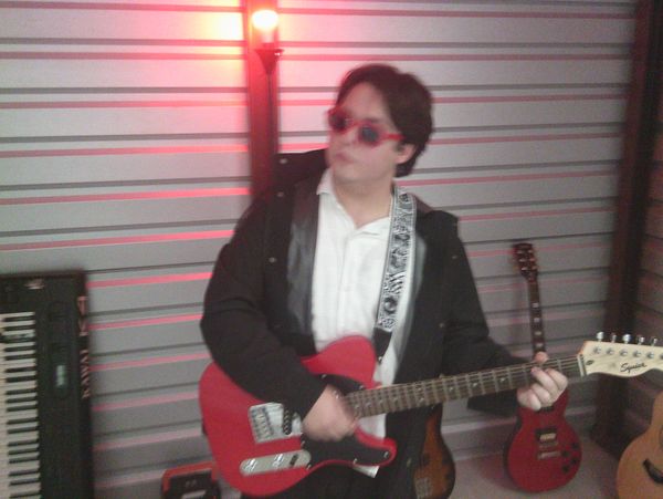 Person playing a red electric guitar with sunglasses indoors.