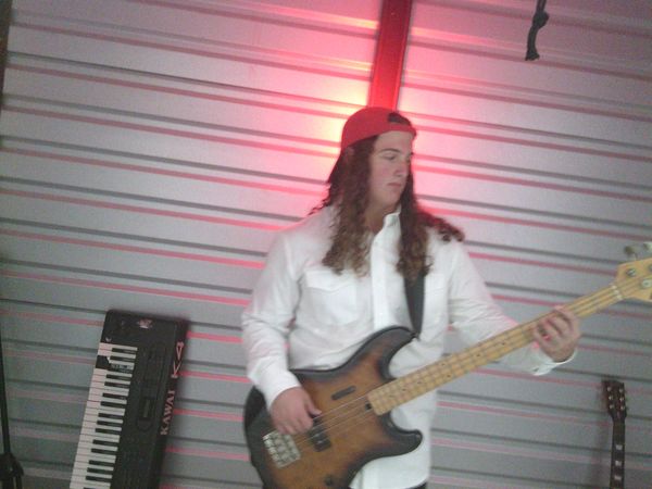Musician in a red cap playing bass guitar with keyboard in background.