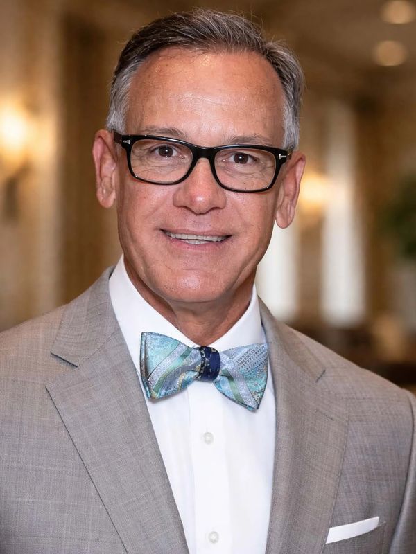 A smiling man in a gray suit with glasses and a colorful bow tie.
