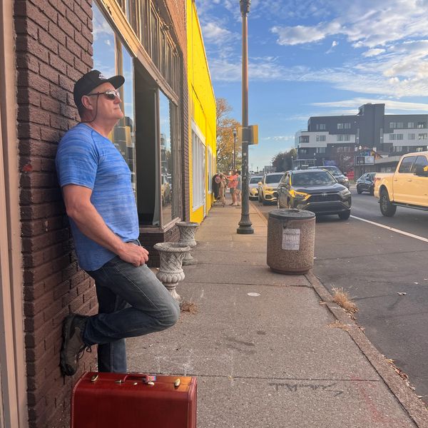 Man in blue shirt leaning on brick wall with suitcase on sidewalk.