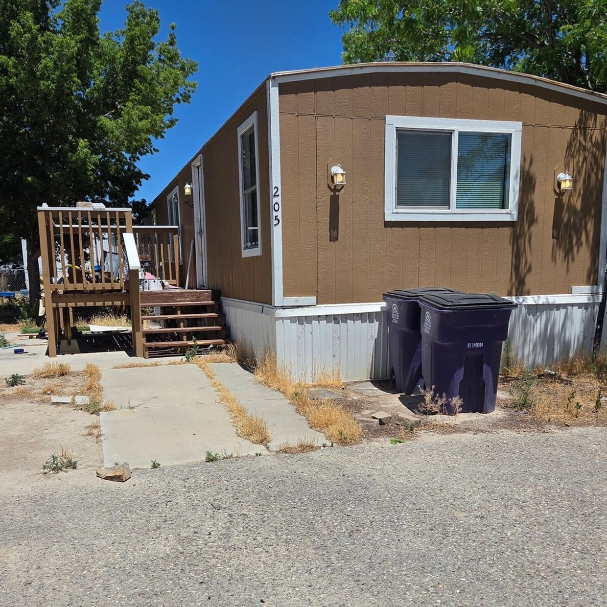 Brown mobile home with stairs and two trash bins outside on a sunny day.