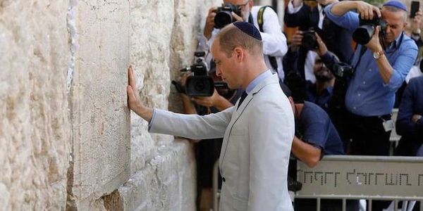 A man in a light suit prays at the Western Wall, surrounded by photographers.