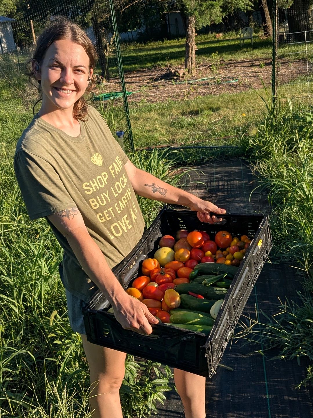 Smiling woman holding a crate of fresh vegetables in a garden.
