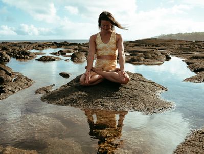 A woman meditates on a rock surrounded by water under a bright sky.