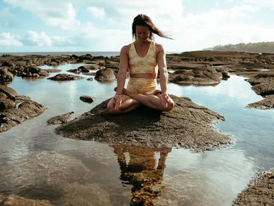 A woman meditates on a rock surrounded by water under a bright sky.