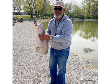 Man holding a large fish by a lakeside on a sunny day.