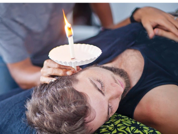 A man lying on a bed with an ear candling candle burning being held by another person.