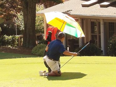 Person kneeling on a golf course holding a club under a colorful umbrella.