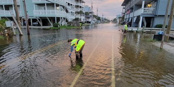 Two people in safety vests inspect a flooded street in a residential area.